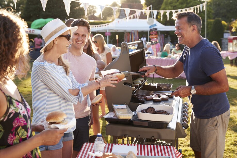 Auf dem Bild sieht man Menschen, die an einem Grill stehen. Ein Man grillt und reicht einer Frau eine Bratwurst.